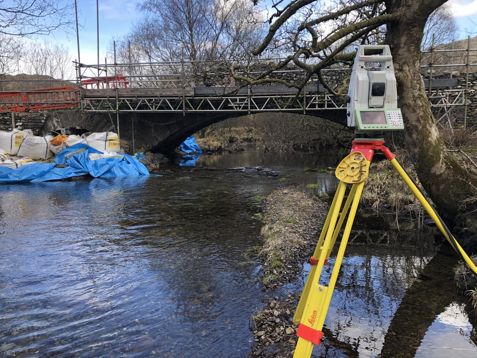 A robotic total station on a river bank, set up facing a scaffolded stone arch bridge during repairs.