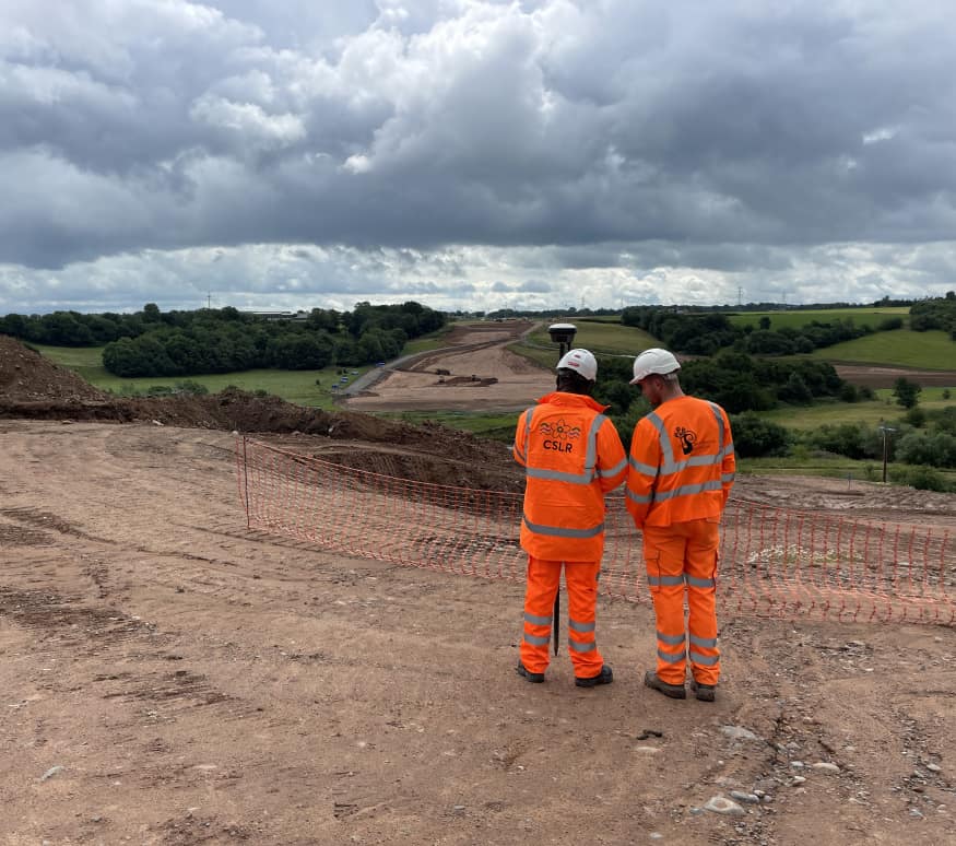Two engineers in orange high-visibility clothing on site, one wearing a Peacock-branded jacket.