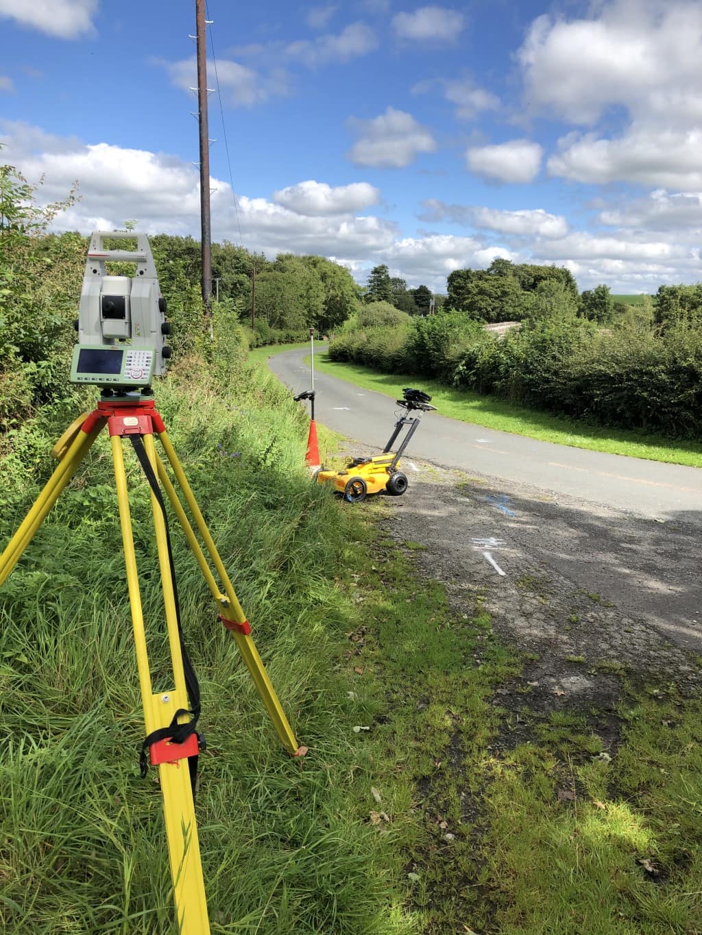 A robotic total station on a verge and a yellow GPR cart on a rural lane, with utility marks painted on the tarmac.