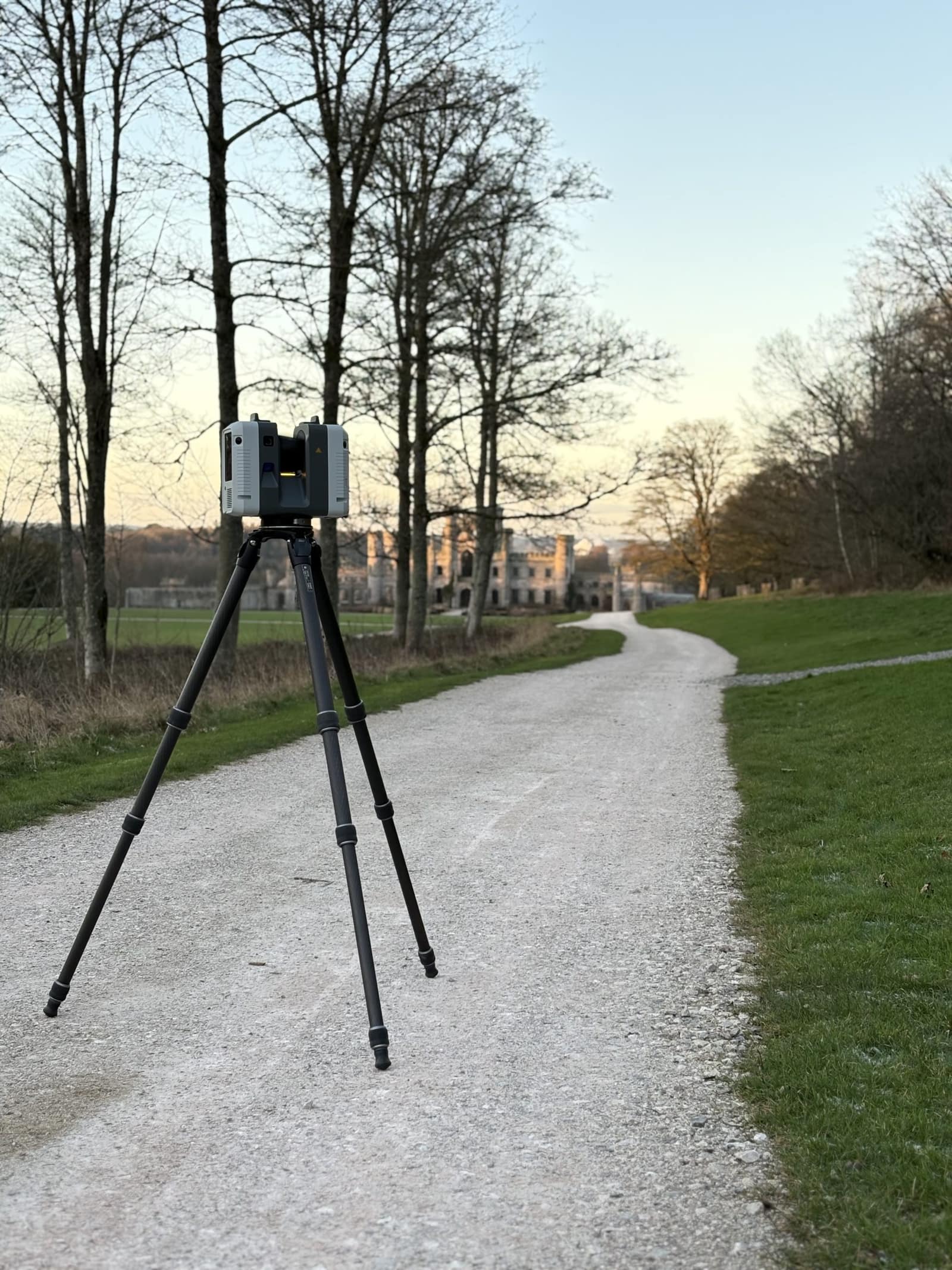 A Leica laser scanner on a carbon tripod on an estate drive, with a castle in the distance behind it.