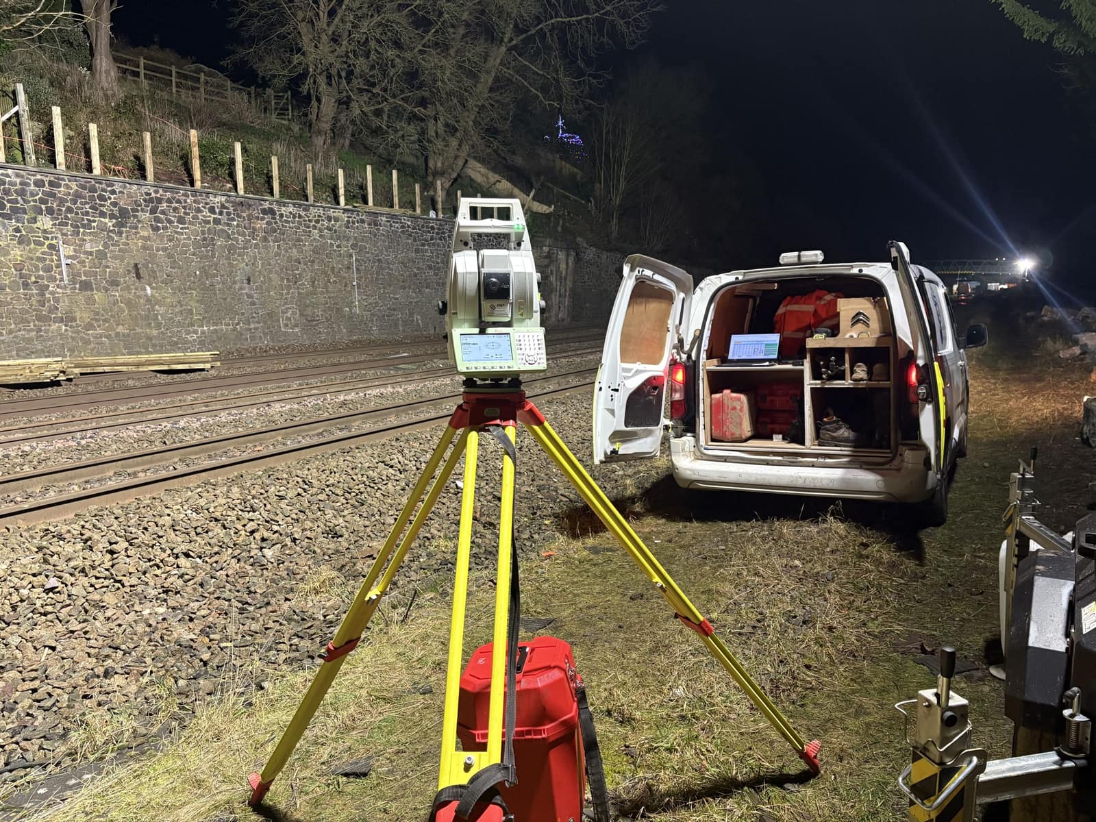 A robotic total station set up beside the track at night during a rail possession, with a survey van and floodlit works behind.